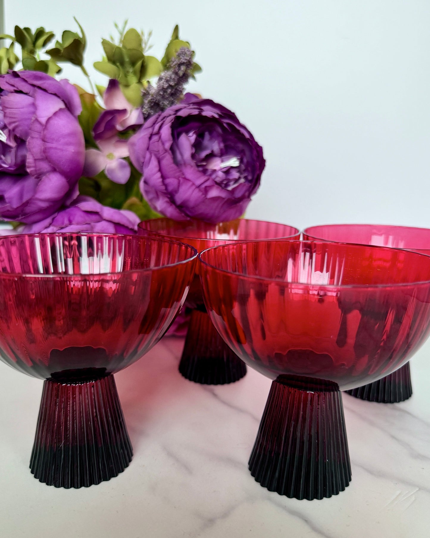 A set of cherry red coupe glass stemware with a dark cherry stem and a brighter cherry-red cup, displayed on a marble surface with purple and green floral decor in the background.