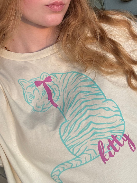 woman wearing a cropped shirt in natural color with a colorful, turquoise blue tiger graphic. The tiger is wearing a pink, coquette bow. The word "kitty" is in pink lettering underneath the tiger. The top is a boxy, cropped t-shirt.
